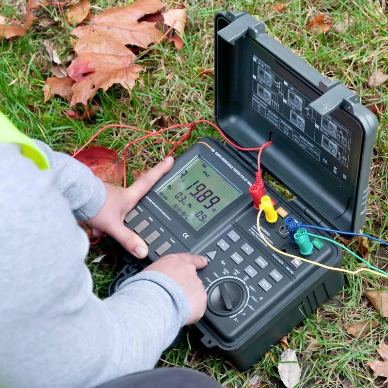 Eine Person nutzt ein digitales Multimeter im Freien auf Gras mit Herbstblättern. Das Gerät zeigt eine Spannung von 19,98 V an.