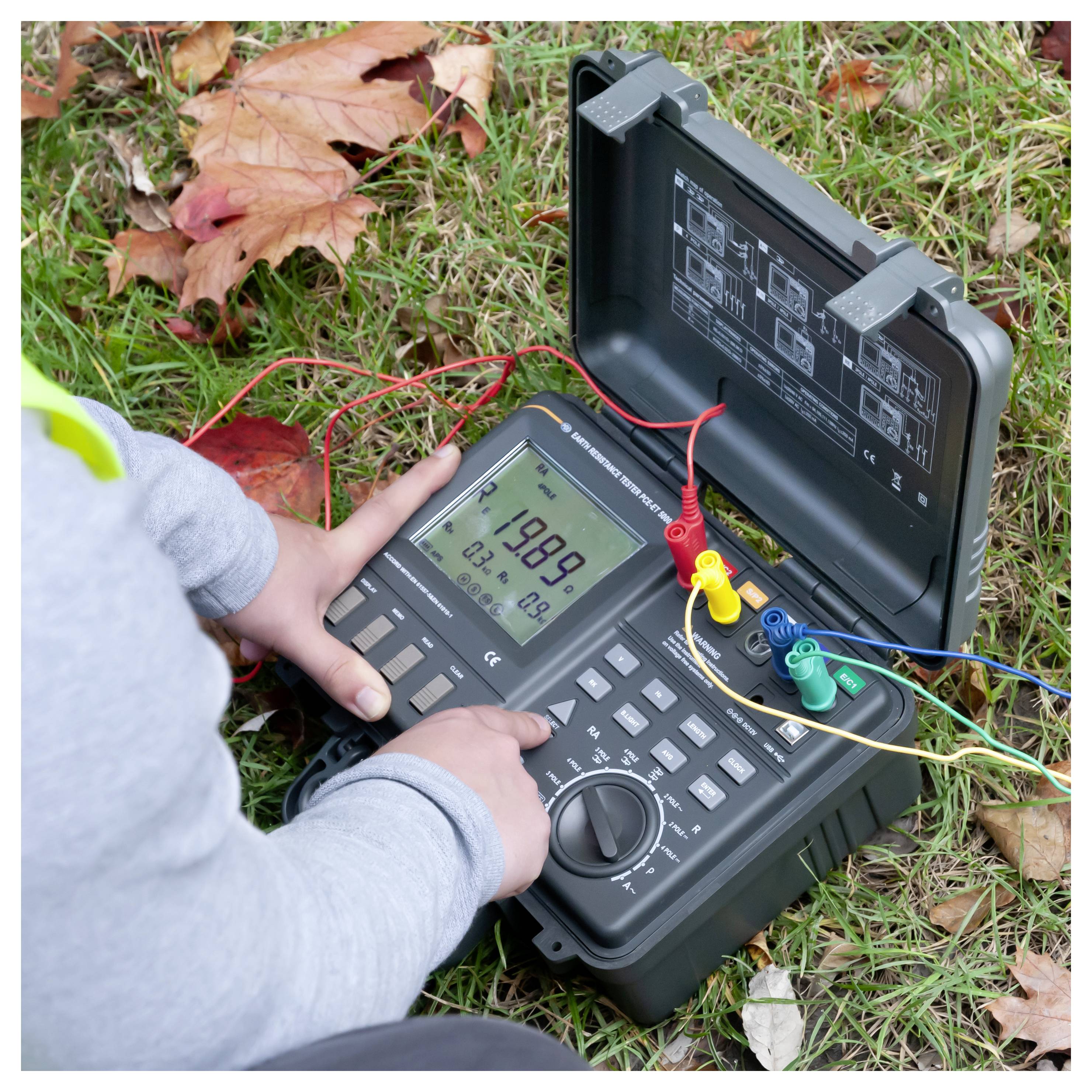 Person, die ein tragbares elektrisches Prüfgerät im Freien auf Gras verwendet, mit Messspitzen, die mit Kabeln verbunden sind, und einem Kofferdeckel, auf dem Schaltpläne zu sehen sind.