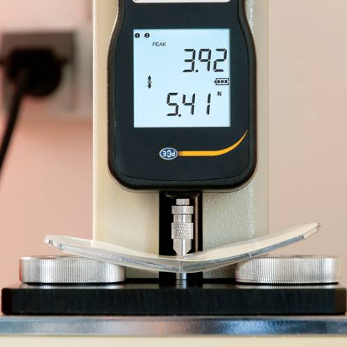 Ein Messgerät mit digitaler Anzeige zeigt 'PEAK 3.92' und '5.41 N'. Es misst den Druck auf ein leicht gebogenes Objekt zwischen zwei Scheiben.