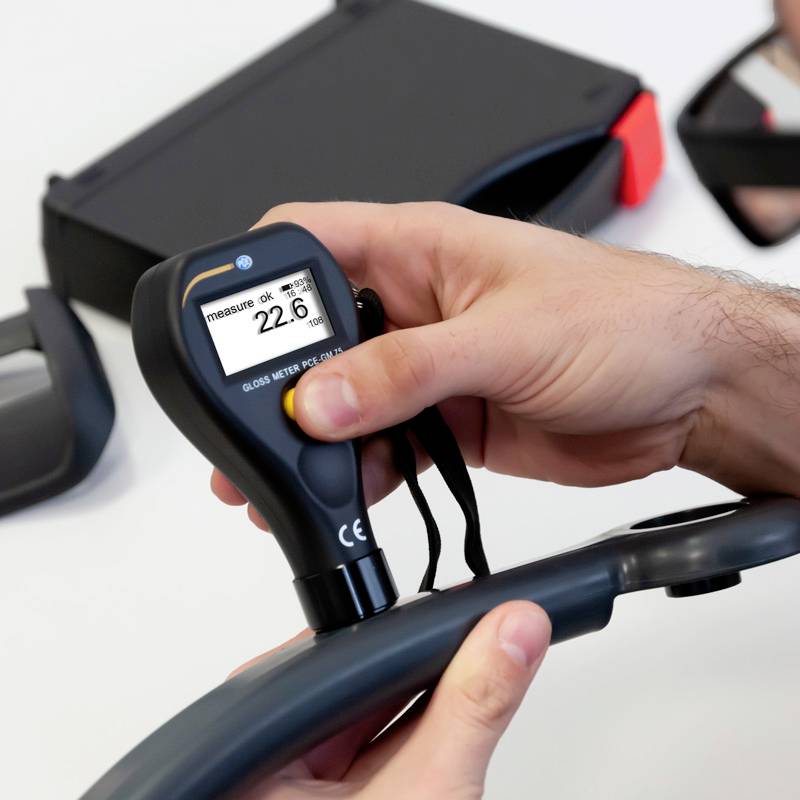 Eine Person hält ein digitales Messgerät, das 'measure ok 22.6 µm' anzeigt, für die Lackdicke eines Autoteils. Im Hintergrund ein Koffer.