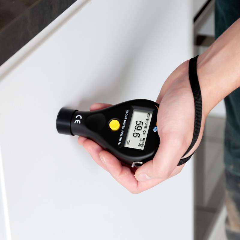 Eine Hand hält ein Gerät zur Messung von Glanz, das auf eine weiße Oberfläche gerichtet ist. Das Gerät zeigt einen Glanzwert von 95,6 an.