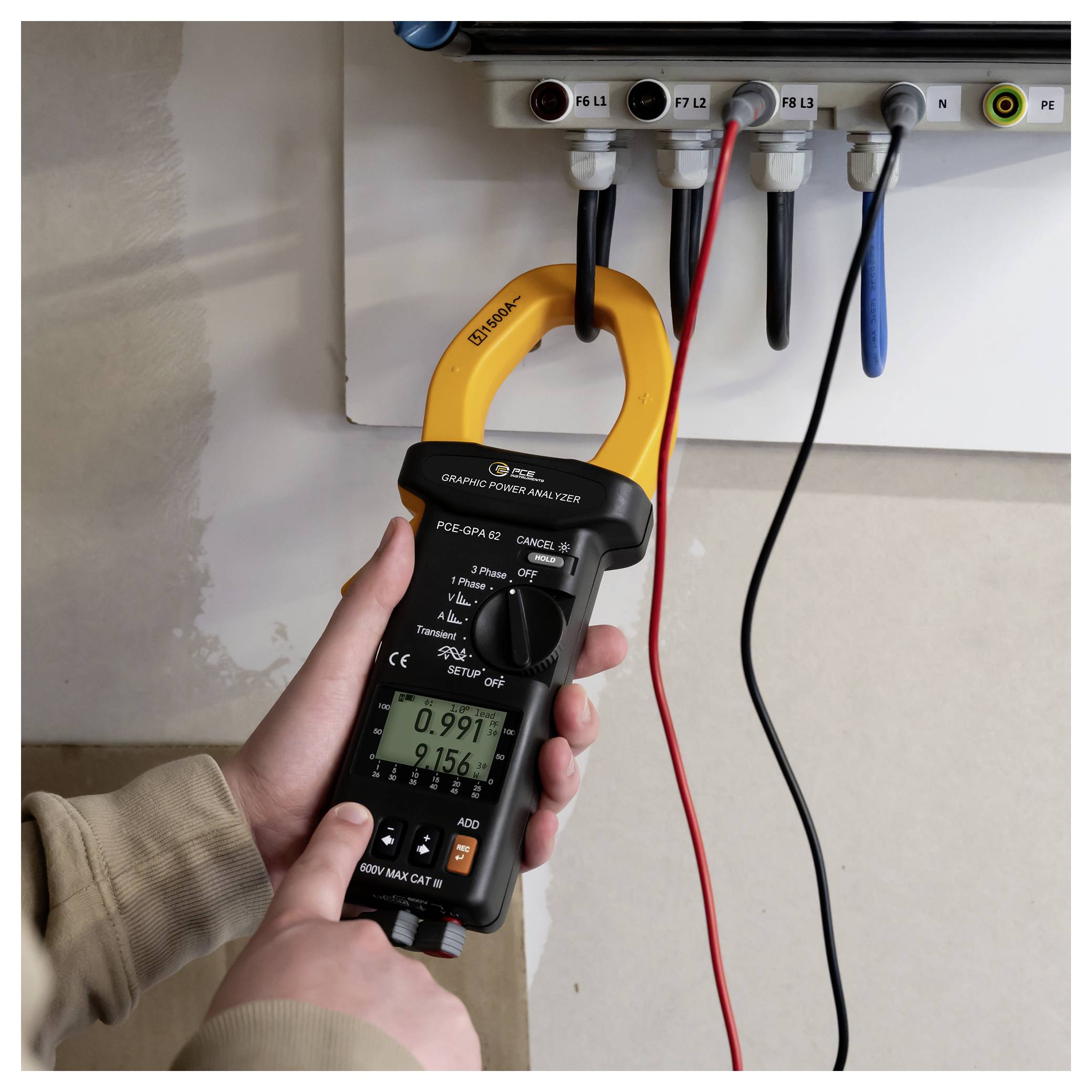 Hand, die eine Zangenstromzange hält und den elektrischen Strom in einem Schaltschrank mit farbigen Drähten und beschrifteten Anschlüssen misst.