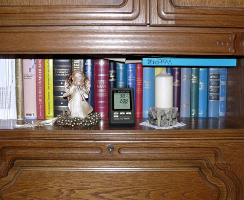 Ein Regal mit Büchern, darunter ein Standbild eines Engels, eine Kerze in einem Halter und ein Thermometer, das 38,1°C und 20,8% anzeigt.