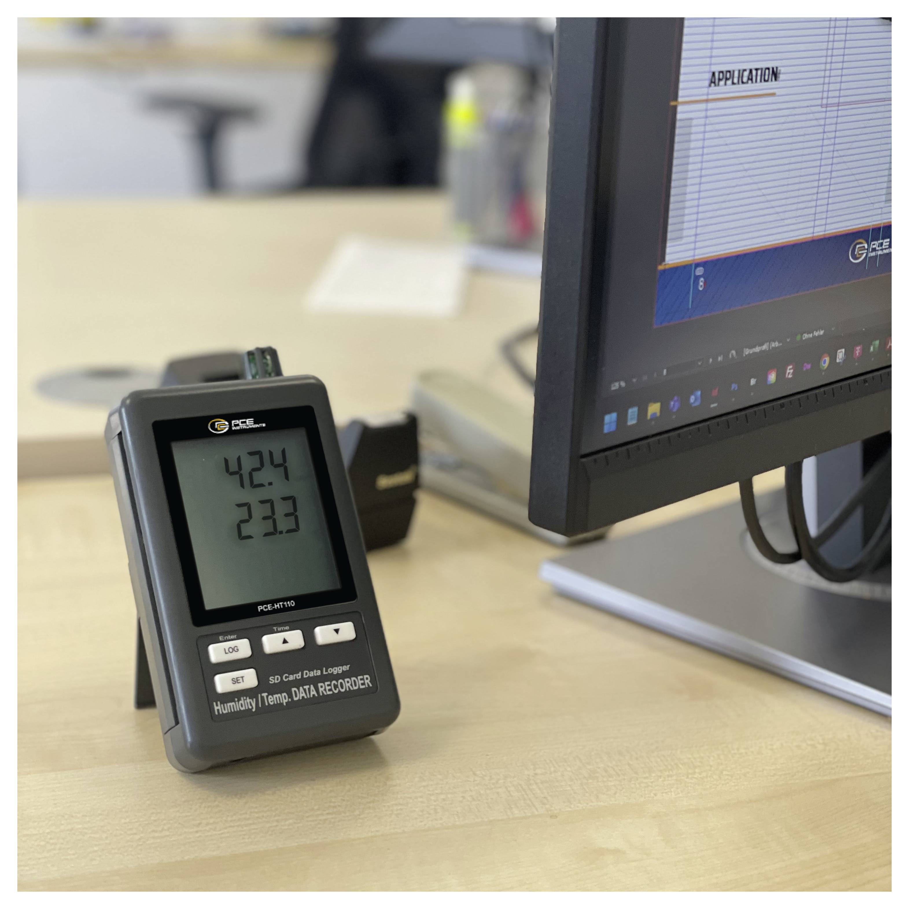 Luftfeuchte- und Temperaturdatenrecorder auf einem Schreibtisch neben einem Computermonitor, der eine Tabellenkalkulationsanwendung anzeigt.