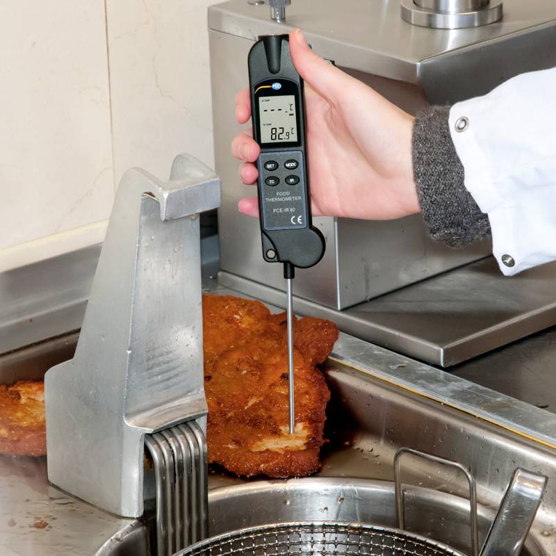 Eine Person misst mit einem digitalen Thermometer die Temperatur von paniertem Fleisch in einem Edelstahlbehälter. Die Anzeige zeigt 82,5 Grad Celsius.