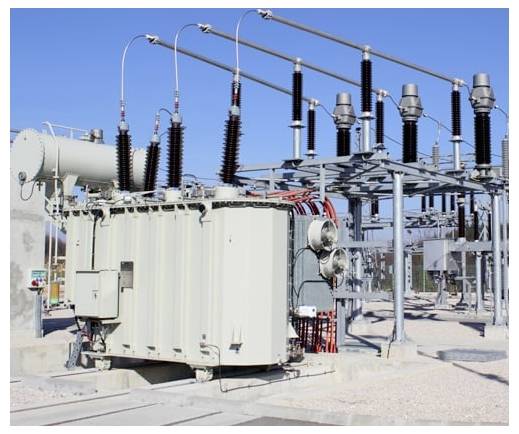 Großer elektrischer Transformator und Hochspannungsumspannwerk-Ausrüstung mit Isolatoren und Stromleitungen im Freien unter klarem blauem Himmel.