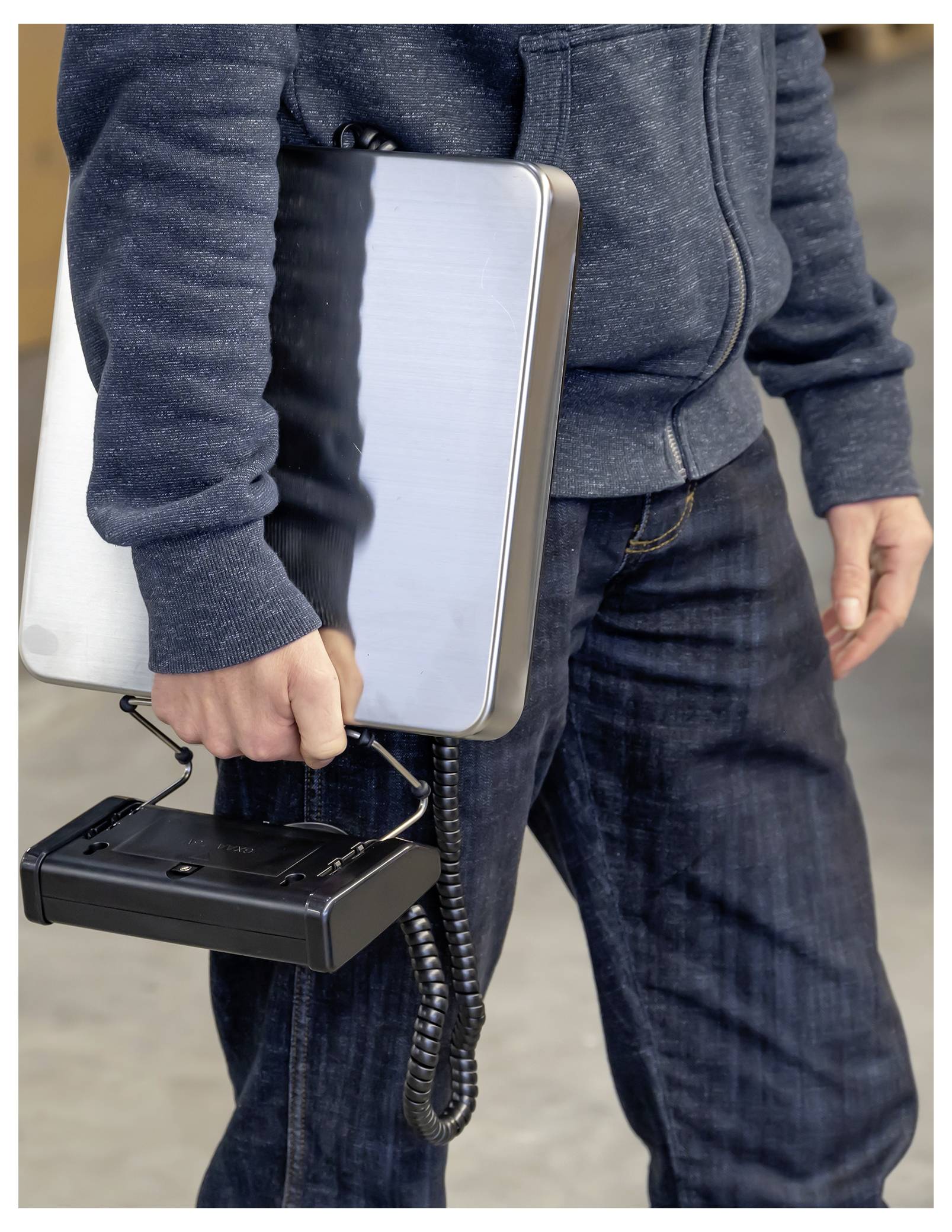 Eine Person hält einen silbernen Laptop unter einem Arm und einen schwarzen schnurlosen Telefonsteckdosen in der anderen Hand, was darauf hindeutet, dass Ausrüstung getragen oder bewegt wird.