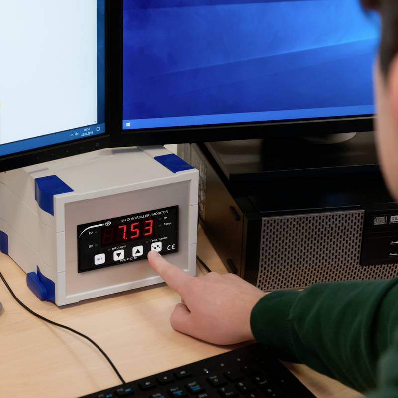 Eine Hand zeigt auf einen pH-Monitor mit der Anzeige '7.53'. Monitore im Hintergrund. Das Bild zeigt eine Laborumgebung.