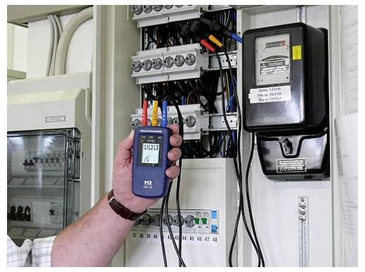Eine Hand hält ein elektrisches Messinstrument vor einem offenen Stromzähler und einer Schaltafeltafel und zeigt eine Stromprüfung oder Messung.