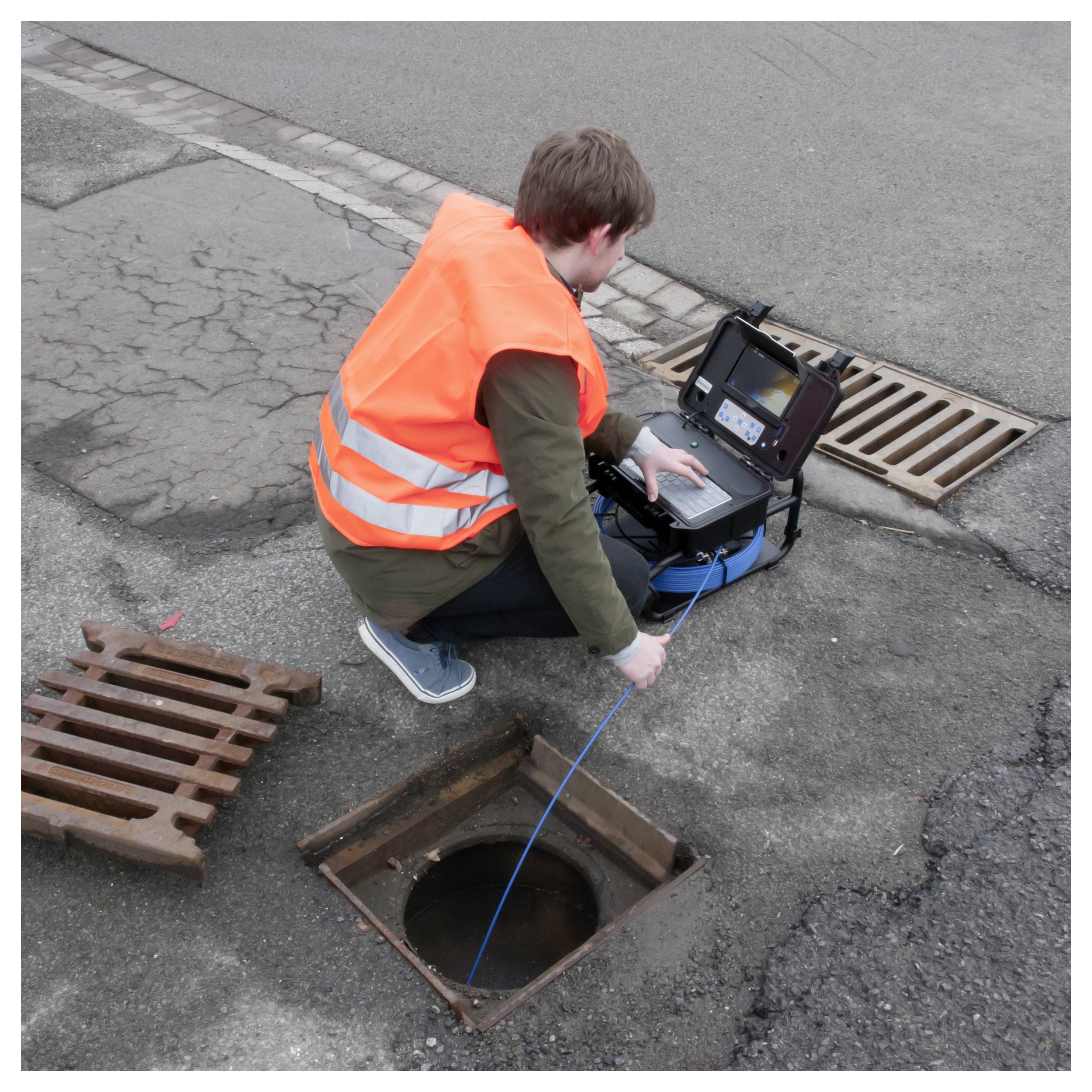 Arbeiter in orangefarbener Sicherheitsweste, der ein Kabelprüfgerät verwendet, um eine offene Straßenschachtöffnung zu untersuchen.