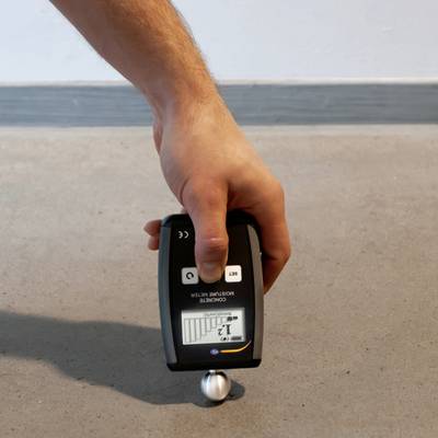 Hand hält ein Messgerät zur Feuchtigkeitsmessung auf einem Betonboden. Das Display zeigt einen Balkenwert an.