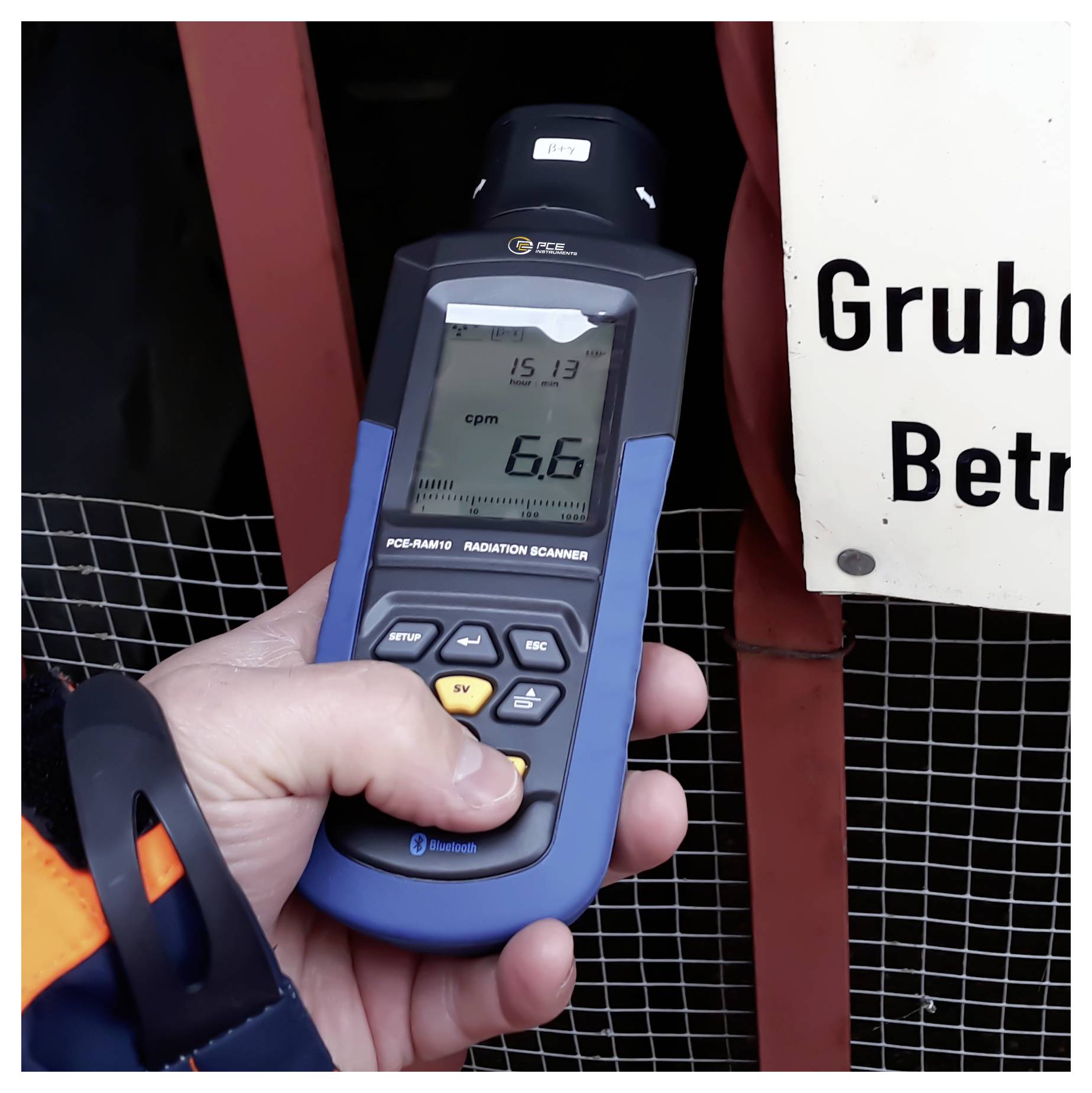 Tragbarer Strahlungsdetektor, der 6,6 cpm anzeigt, gehalten in der Nähe einer Barriere mit einem sichtbaren Warnschild "Grub...".
