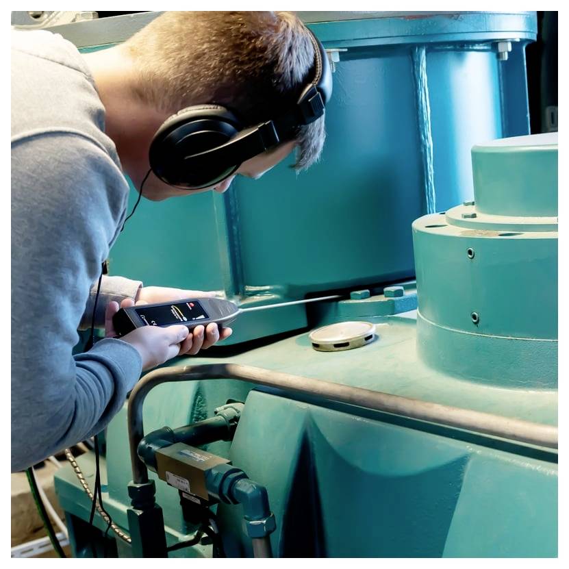 Mann mit Kopfhörern nutzt ein Smartphone, um türkisfarbene Industriemaschinen in einer Werkstatt oder einem Labor zu inspizieren.