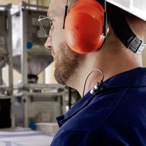 Eine Person mit rotem Gehörschutz schaut in einer Fabrikumgebung konzentriert nach vorne. Im Hintergrund sind Maschinen sichtbar.