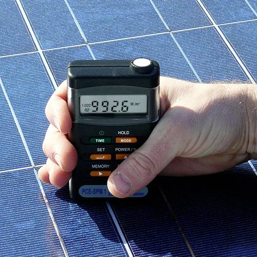 Hand hält ein Gerät zur Messung von Sonnenenergie. Das Display zeigt '992,6 W/m²'. Hintergrund: Solarpanel.