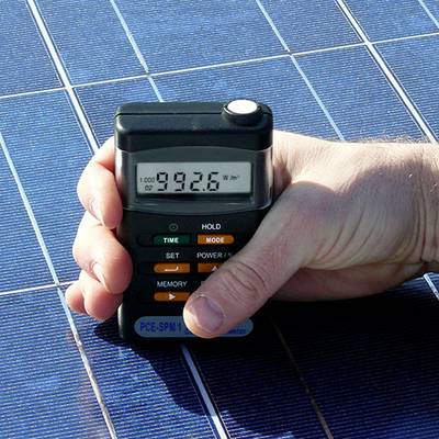 Hand hält ein Gerät zur Messung von Sonnenenergie. Das Display zeigt '992,6 W/m²'. Hintergrund: Solarpanel.