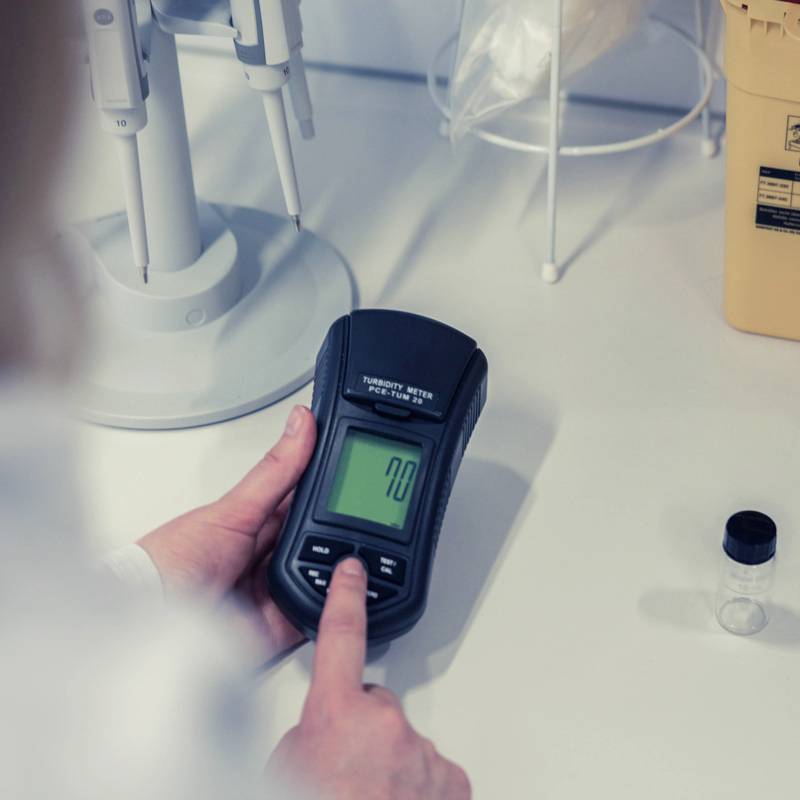 Ein Hand hält ein Messgerät mit digitaler Anzeige, das den Wert '70' zeigt. Daneben stehen weitere Laborgeräte auf einem Tisch.