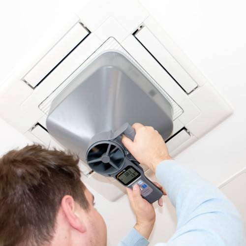 Mann justiert mit einem Messgerät die Belüftungseinheit in der Decke. Das Display zeigt die Temperatur an. Arbeitsplatzinstallation.