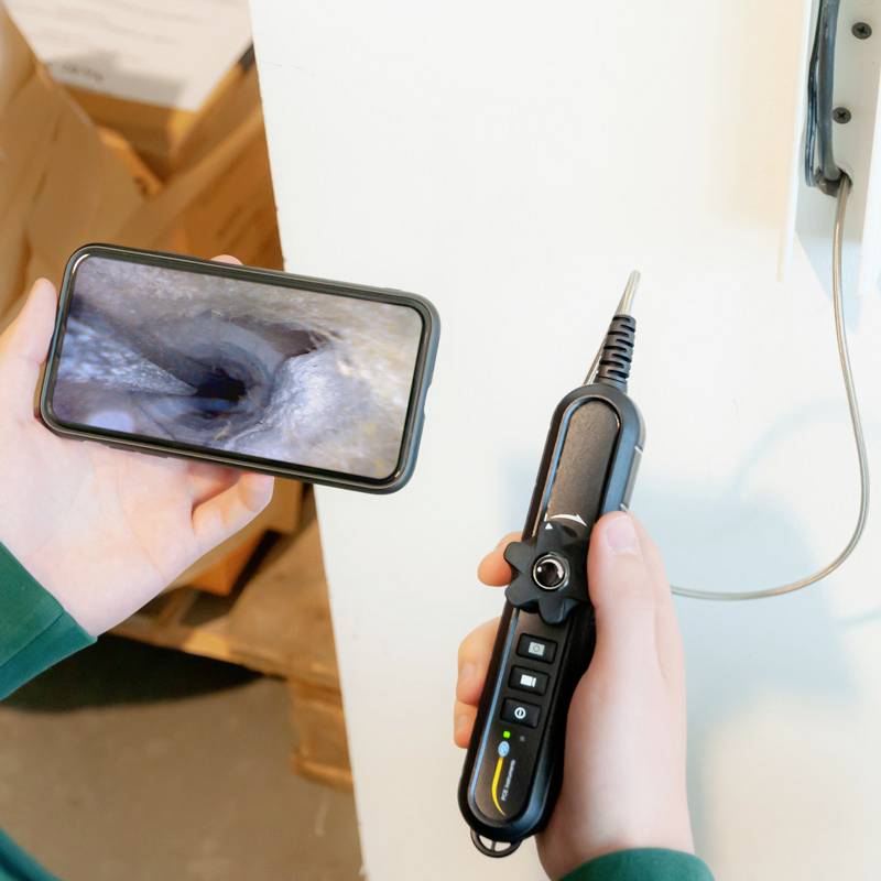 Eine Person hält ein Endoskop und ein Smartphone. Auf dem Handybildschirm ist das Innere eines Rohres zu sehen.