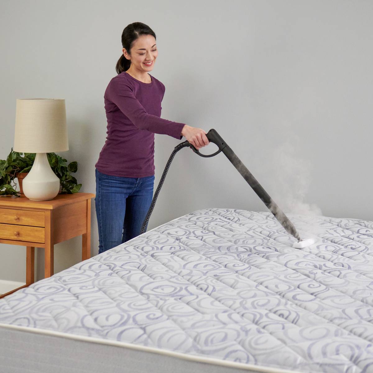 Eine Person reinigt mit einem Dampfreiniger eine Matratze in einem Schlafzimmer. Neben dem Bett steht ein Nachttisch mit einer Lampe.