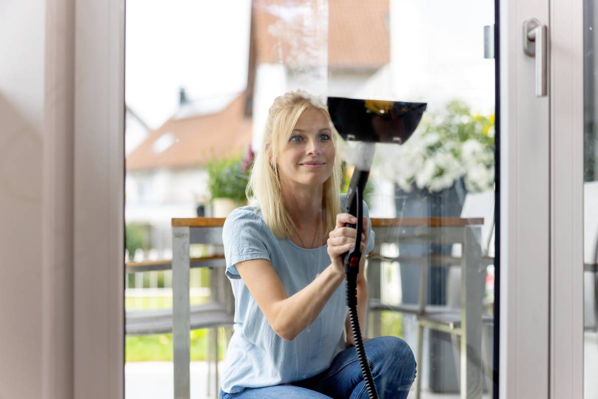 Eine Person reinigt ein Fenster mit einem Dampfreiniger. Im Hintergrund ist ein Teil eines Hauses und Pflanzen zu sehen.