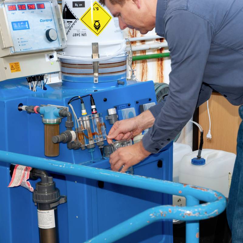 Ein Mann prüft die Einstellungen an einem Wasserfiltrationssystem in einer industriellen Umgebung. Chemikalienbehälter im Hintergrund.