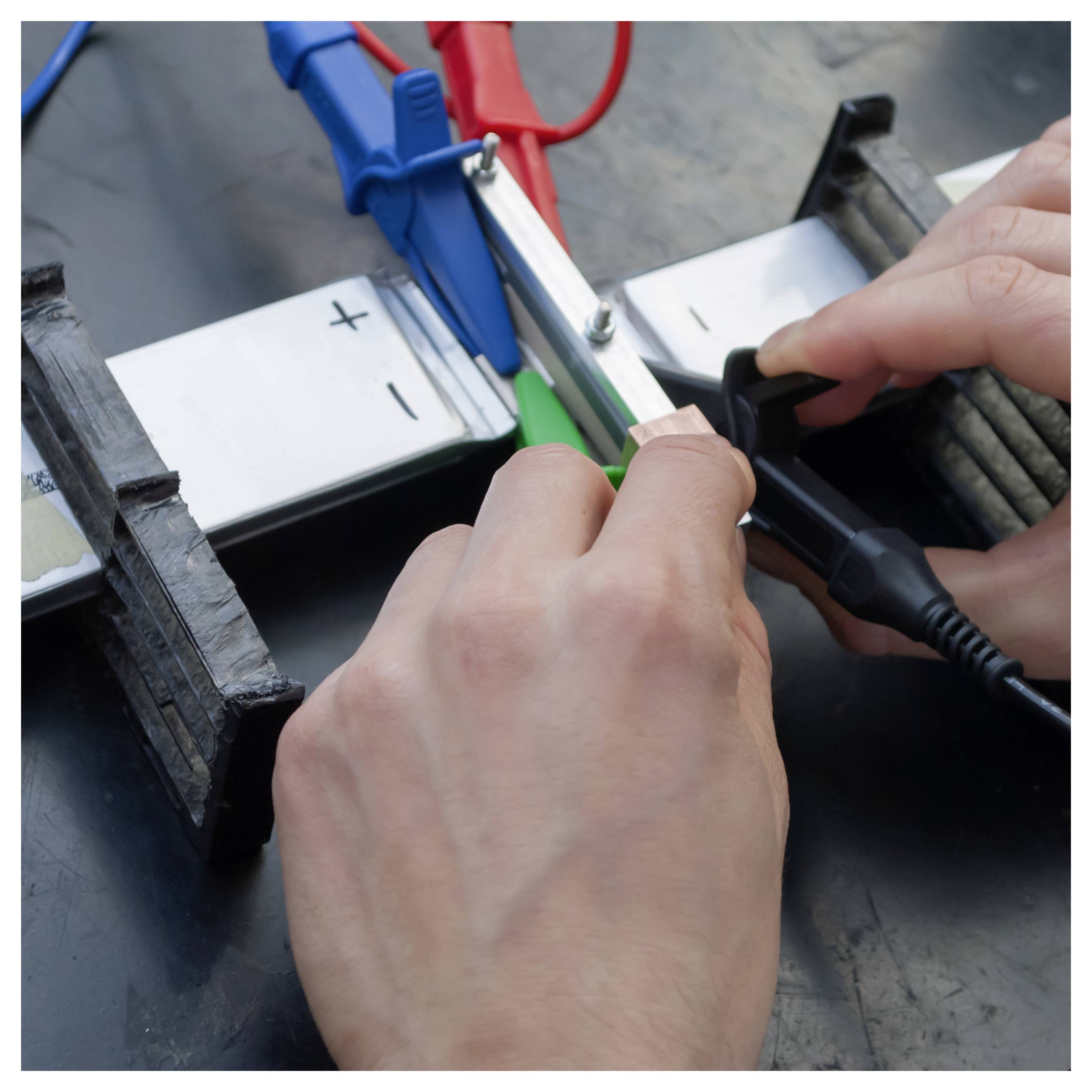 Hände verbinden farbige Messleitungen mit einem mit „+" und „-" gekennzeichneten Batteriepack und verwenden dabei eine Multimeter-Sonde.
