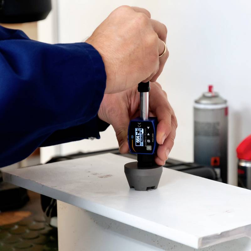 Eine Person in Arbeitskleidung misst die Härte oder Dicke eines weißen Oberflächenmaterials mit einem Messgerät. Hintergrund unscharf.