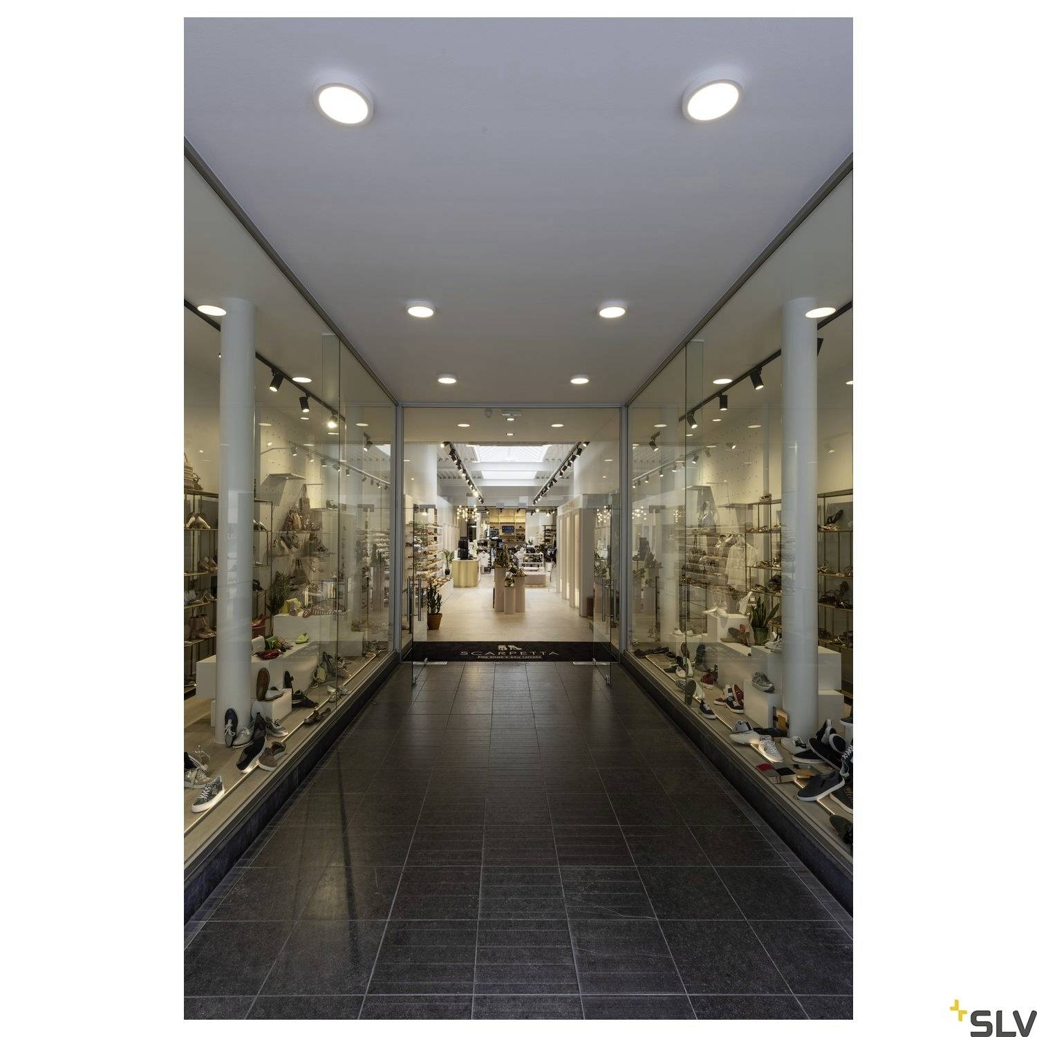 Ein beleuchteter Ladenkorridor mit Glastüren, zeigt modern eingerichtete Innenräume mit Regalen auf beiden Seiten.