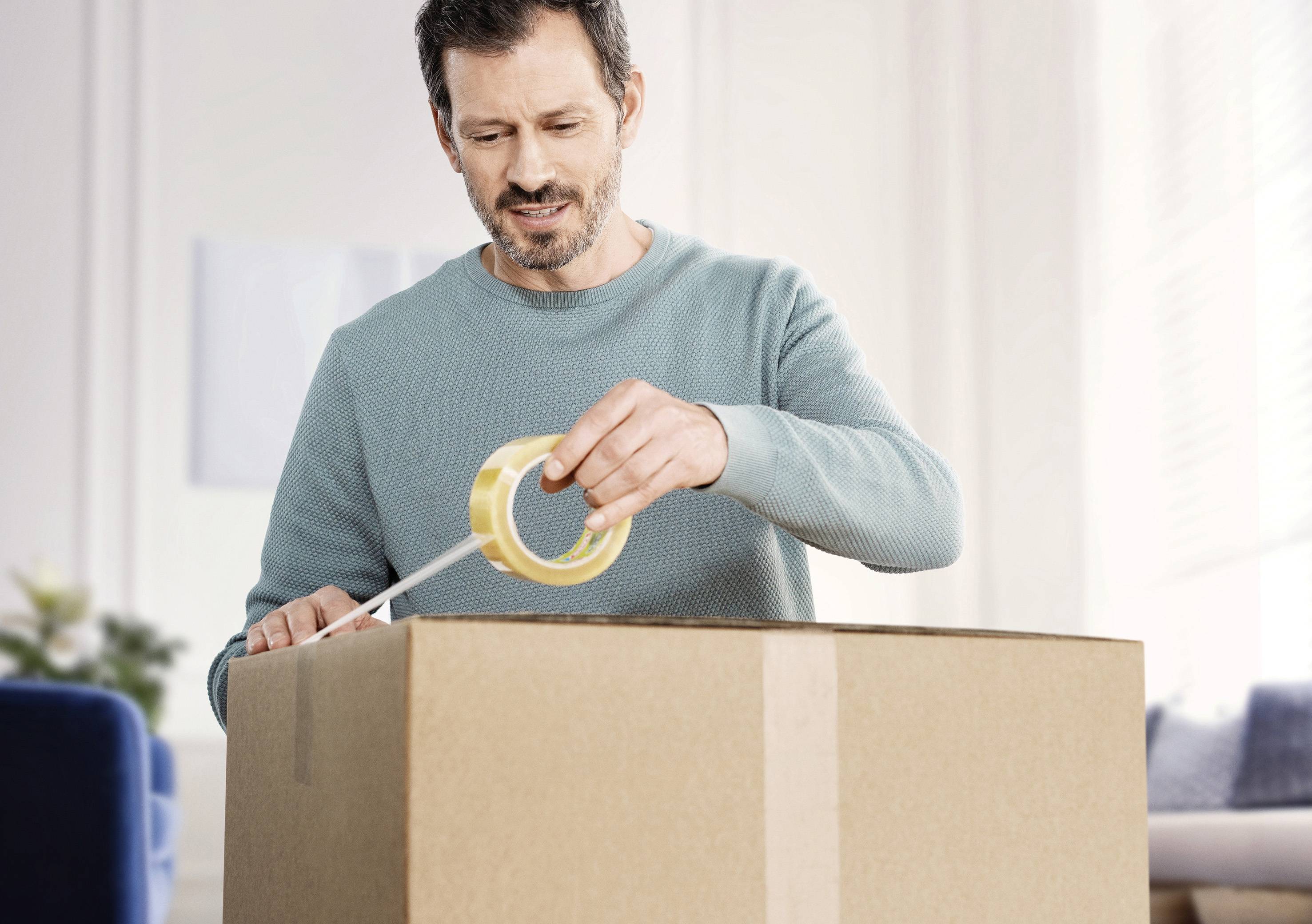 Mann in türkisfarbenem Pullover verschließt einen großen Karton mit Klebeband in einem hellen Raum.
