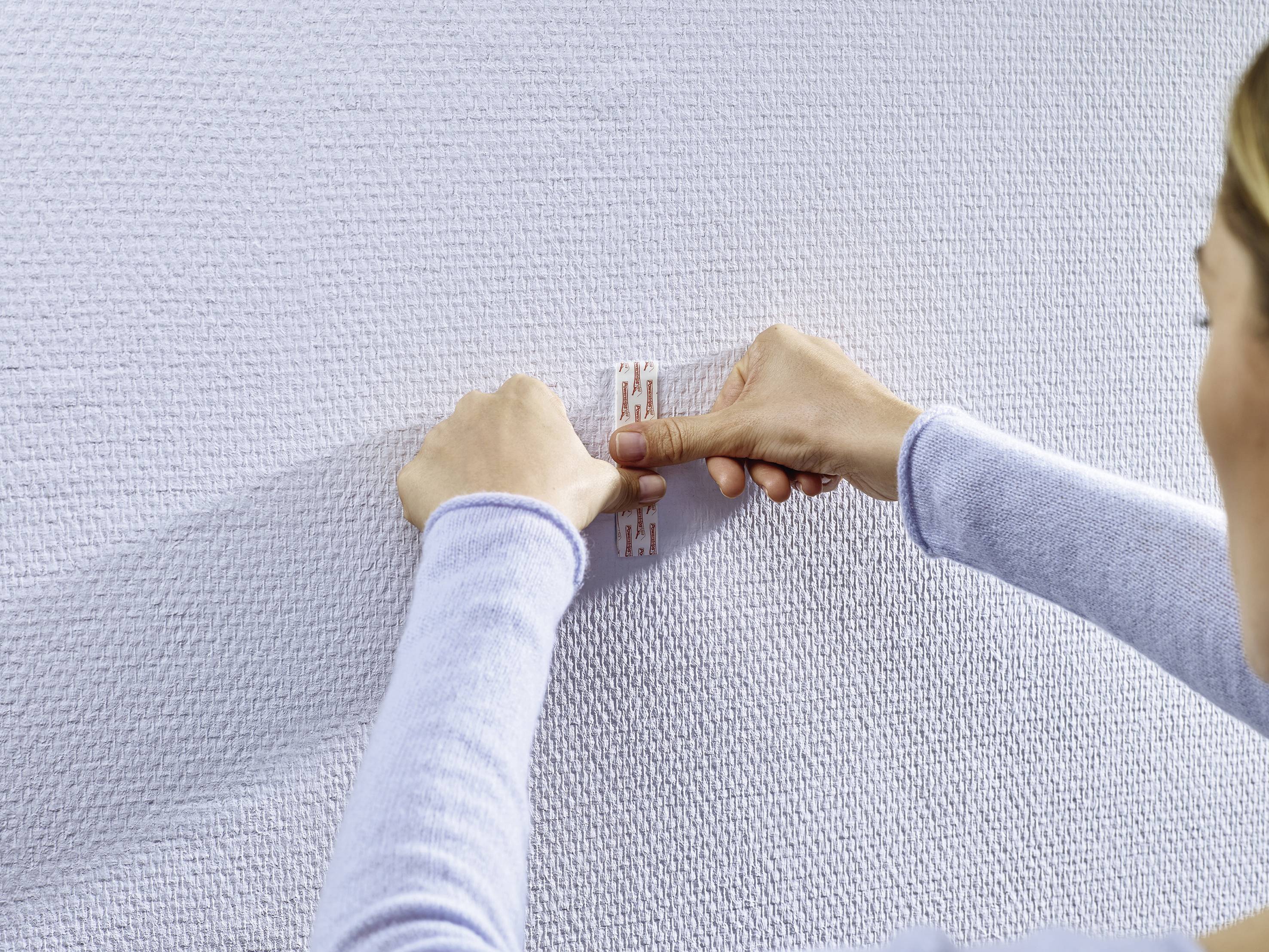 Eine Person befestigt ein kleines, weißes Kunststoffteil an einer strukturierten weißen Wand. Hände und Teil sind im Fokus.