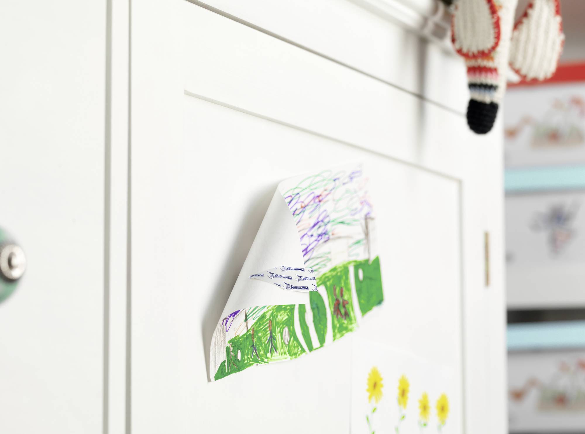 Eine bunte Kinderzeichnung eines Hauses und einer Wiese hängt an einem weißen Schrank. Oben rechts hängt ein gestricktes Stofftier.