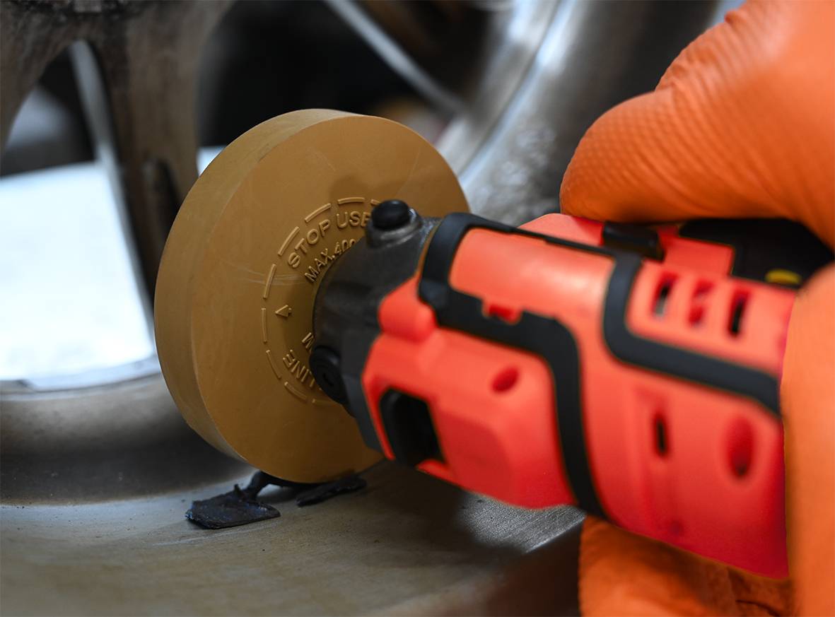 Eine Person entfernt Aufkleber von einer Metalloberfläche mit einem orangefarbenen Werkzeug, das ein rotierendes Rad nutzt.