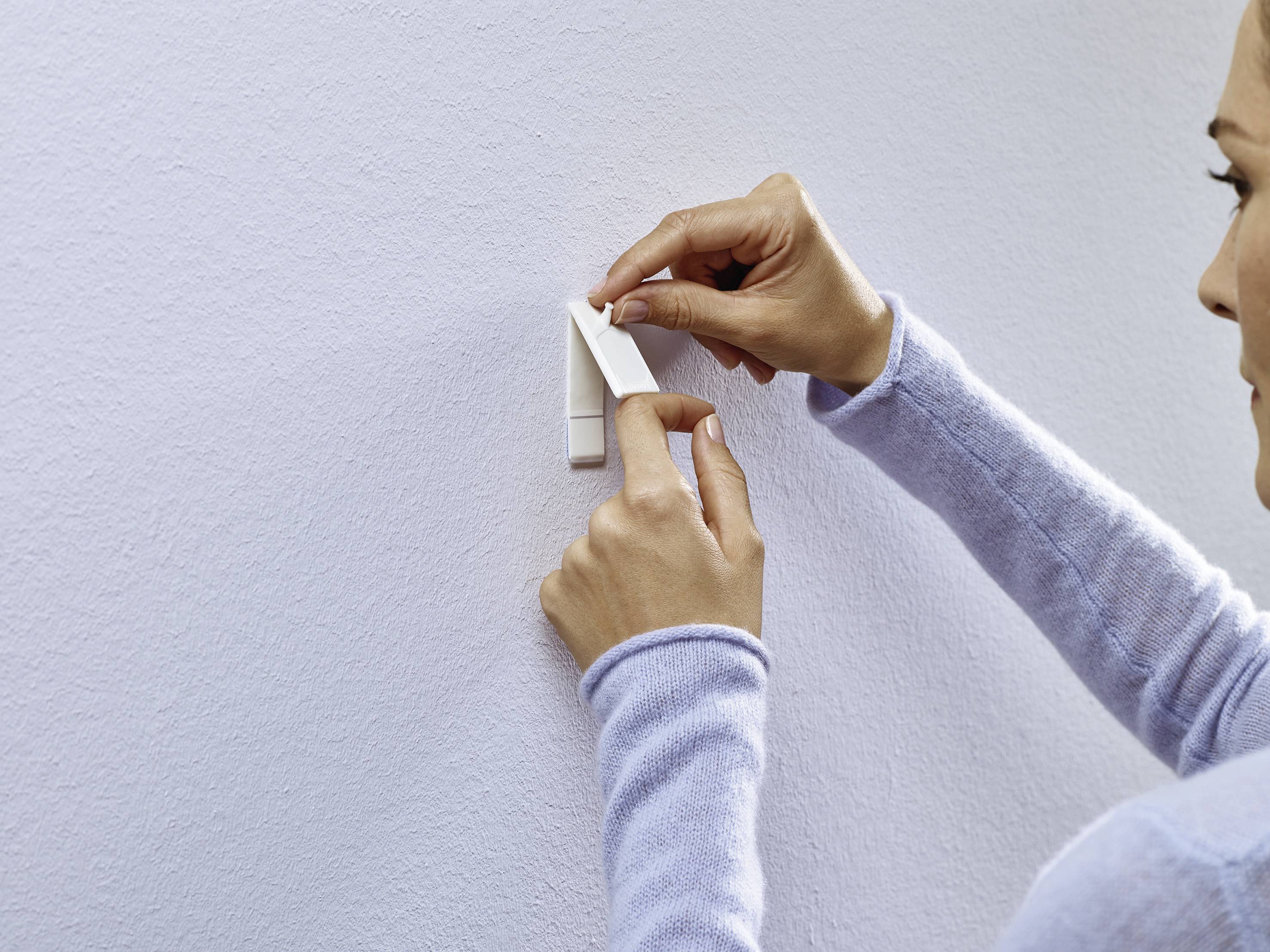Eine Person bringt einen Klebestreifen an einer weißen Wand an. Sie trägt einen hellblauen Pullover.