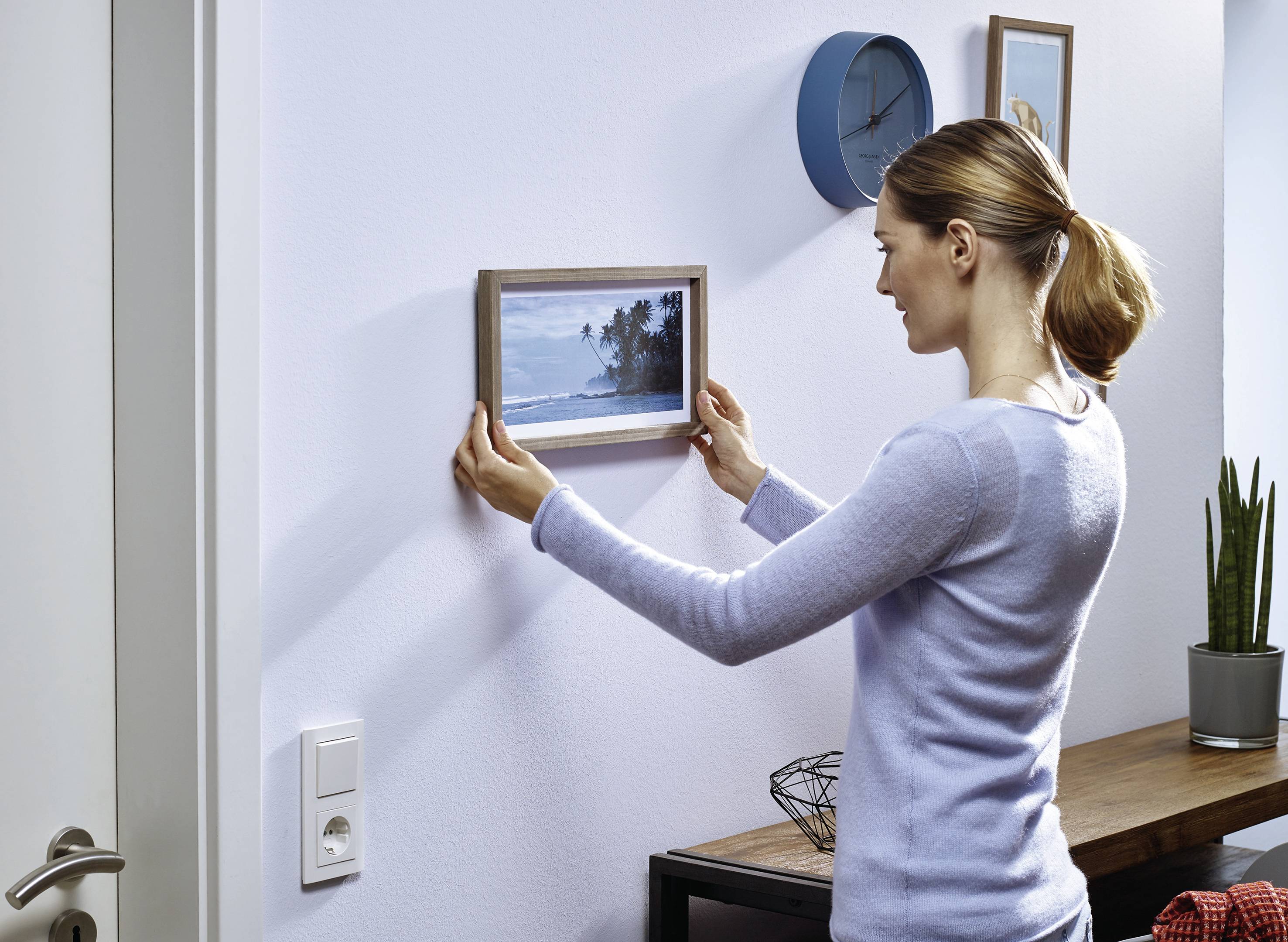 Eine Frau hängt ein gerahmtes Bild mit Strandmotiv an die Wand. Im Hintergrund sind eine Uhr und Pflanzen zu sehen.
