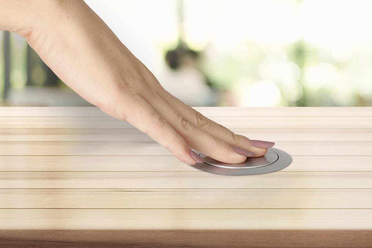 Eine Hand drückt einen runden Knopf in einem Holztisch. Hintergrund ist unscharf, natürliche Beleuchtung erkennbar.