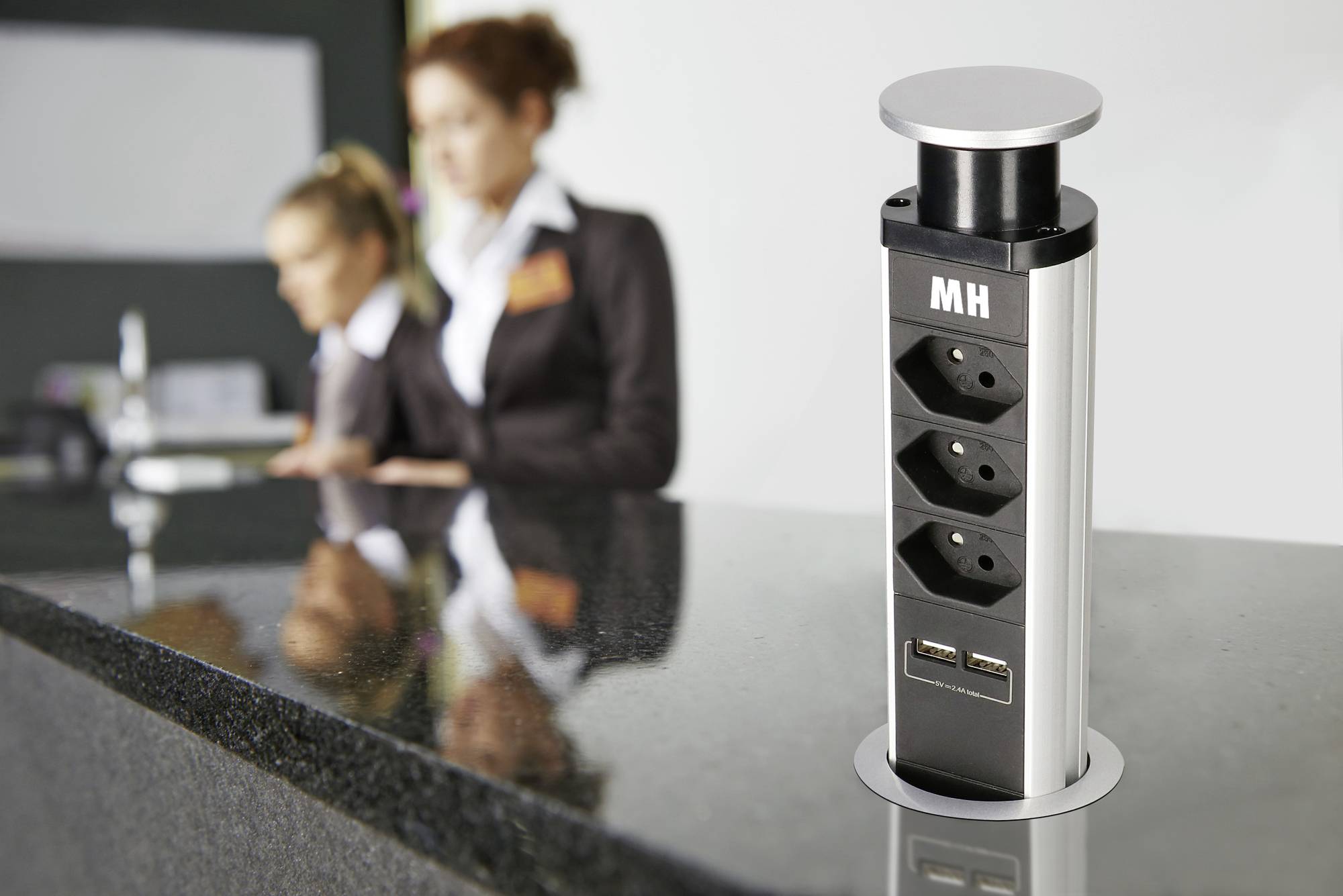 Versenkbare Steckdosenleiste mit zwei USB-Anschlüssen auf einer glänzenden Tischplatte, im Hintergrund zwei unscharfe Personen.