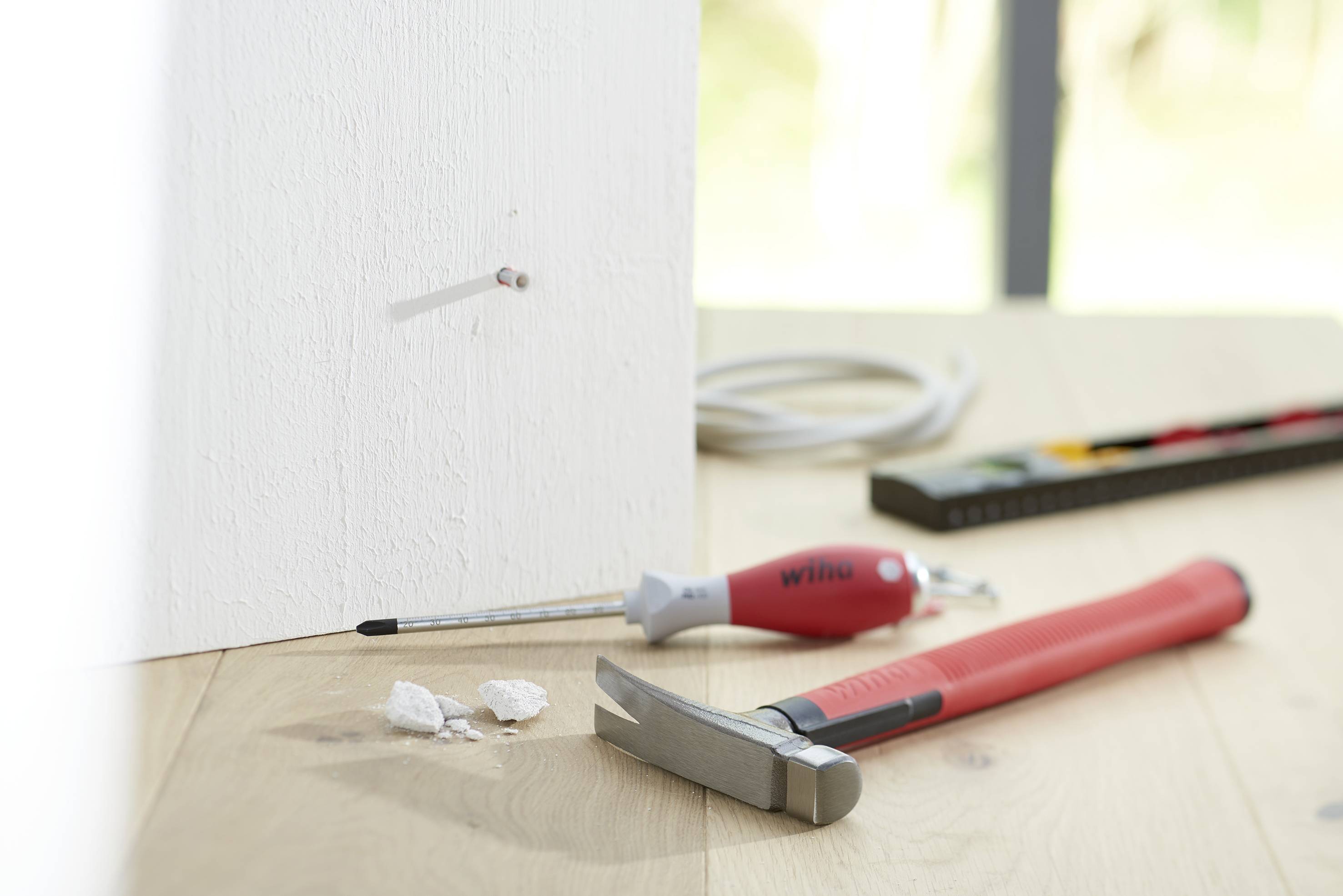 Werkzeugset auf einem Holztisch: Hammer, Schraubendreher und Wasserwaage. Ein Nagel ist schräg in die Wand eingetrieben.