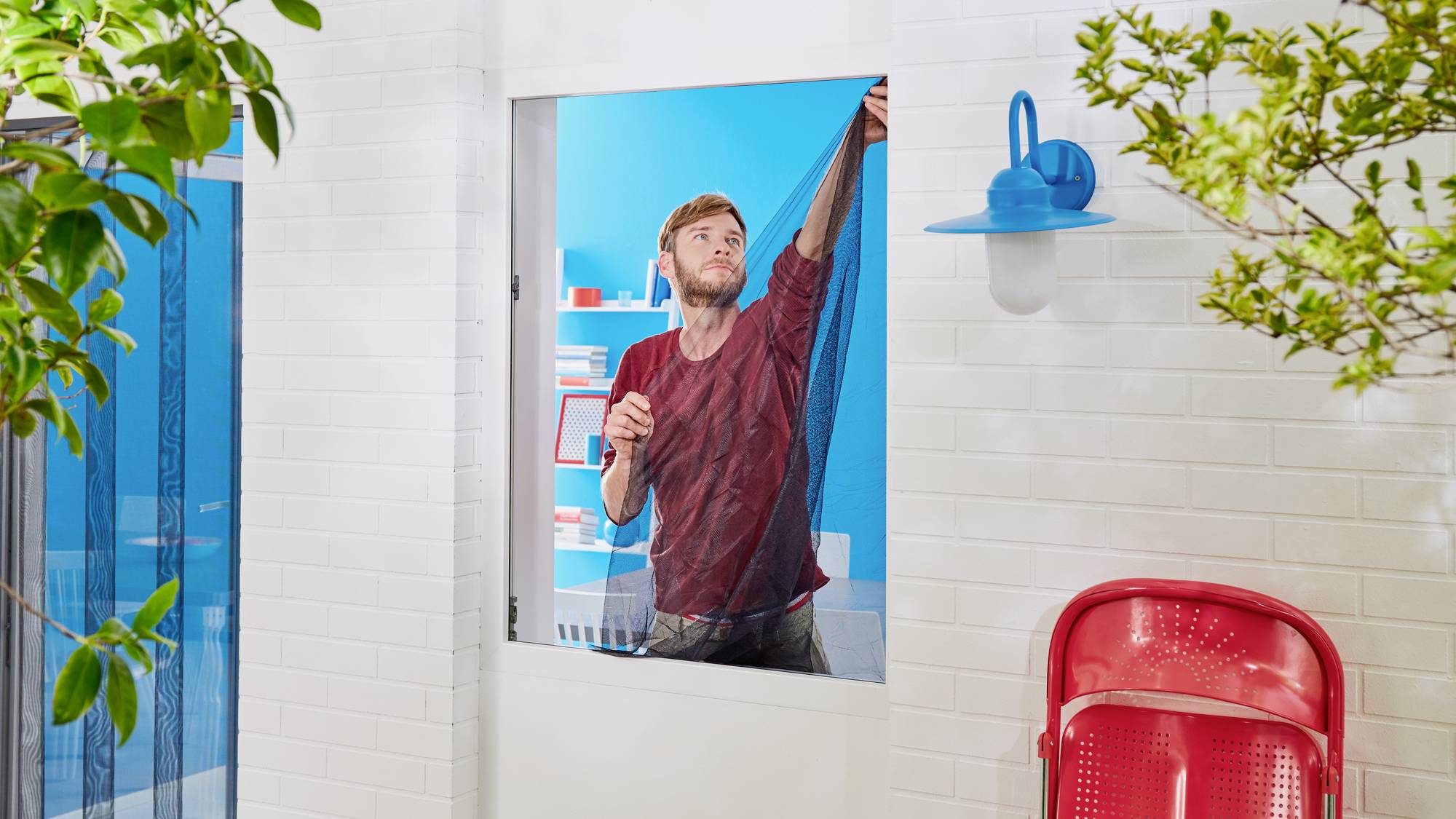 Eine Person streicht eine Fensterleiste blau in einem hellen Raum. Ein roter Stuhl steht draußen an der weißen Ziegelmauer.