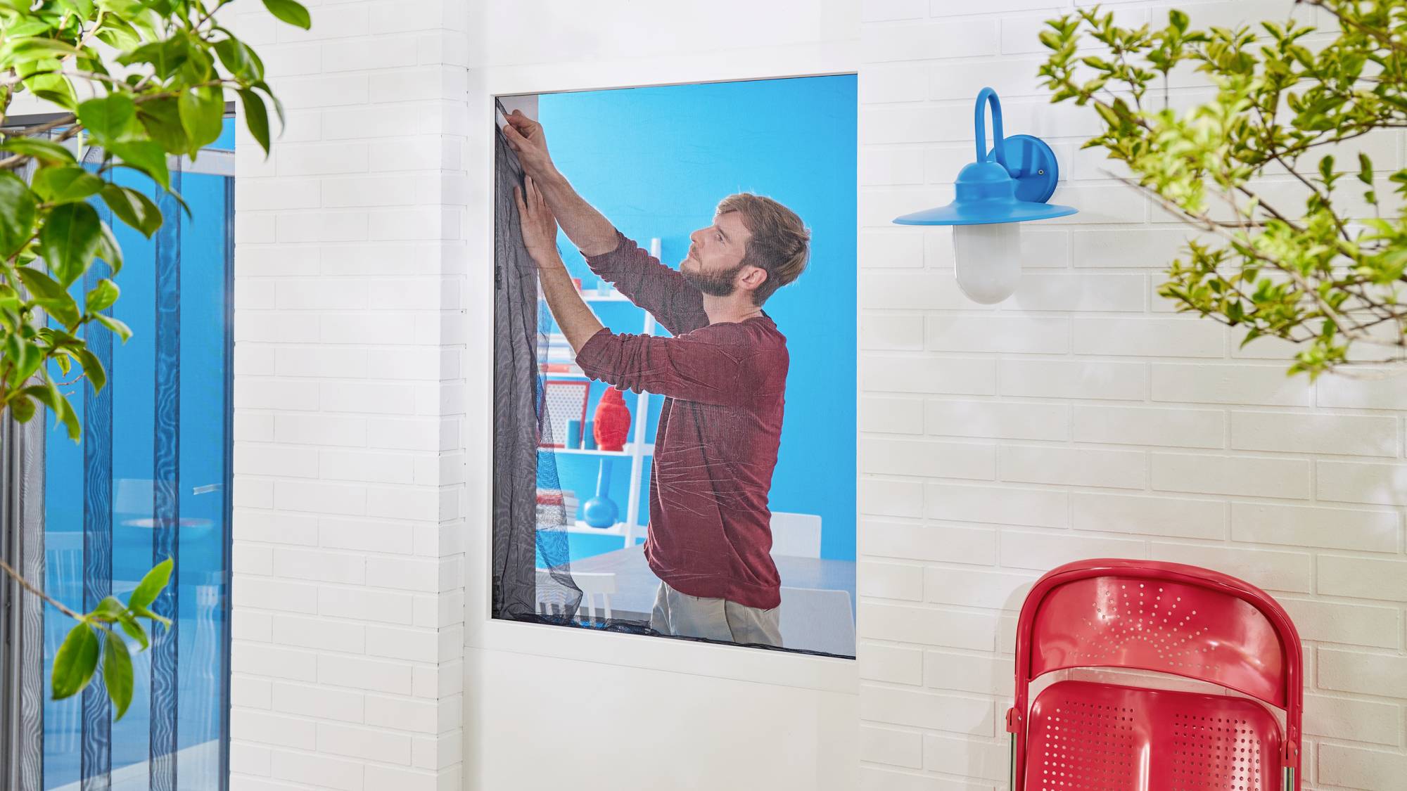 Ein Mann in rotem Pullover zieht dünnen Vorhang vor einem großen Fenster in einem modern gestalteten Raum zu.