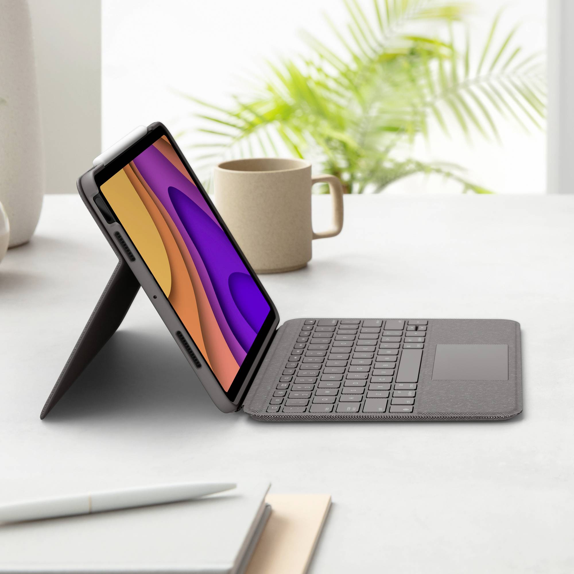 Ein Tablet steht auf einem Schreibtisch in einem Tastatur-Cover, umgeben von einer Tasse und Pflanzen im Hintergrund.