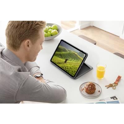 Ein Mann betrachtet ein Tablet mit einem Bild von grünen Feldern. Auf dem Tisch steht ein Glas Orangensaft, ein Gebäck und ein Schlüsselbund.