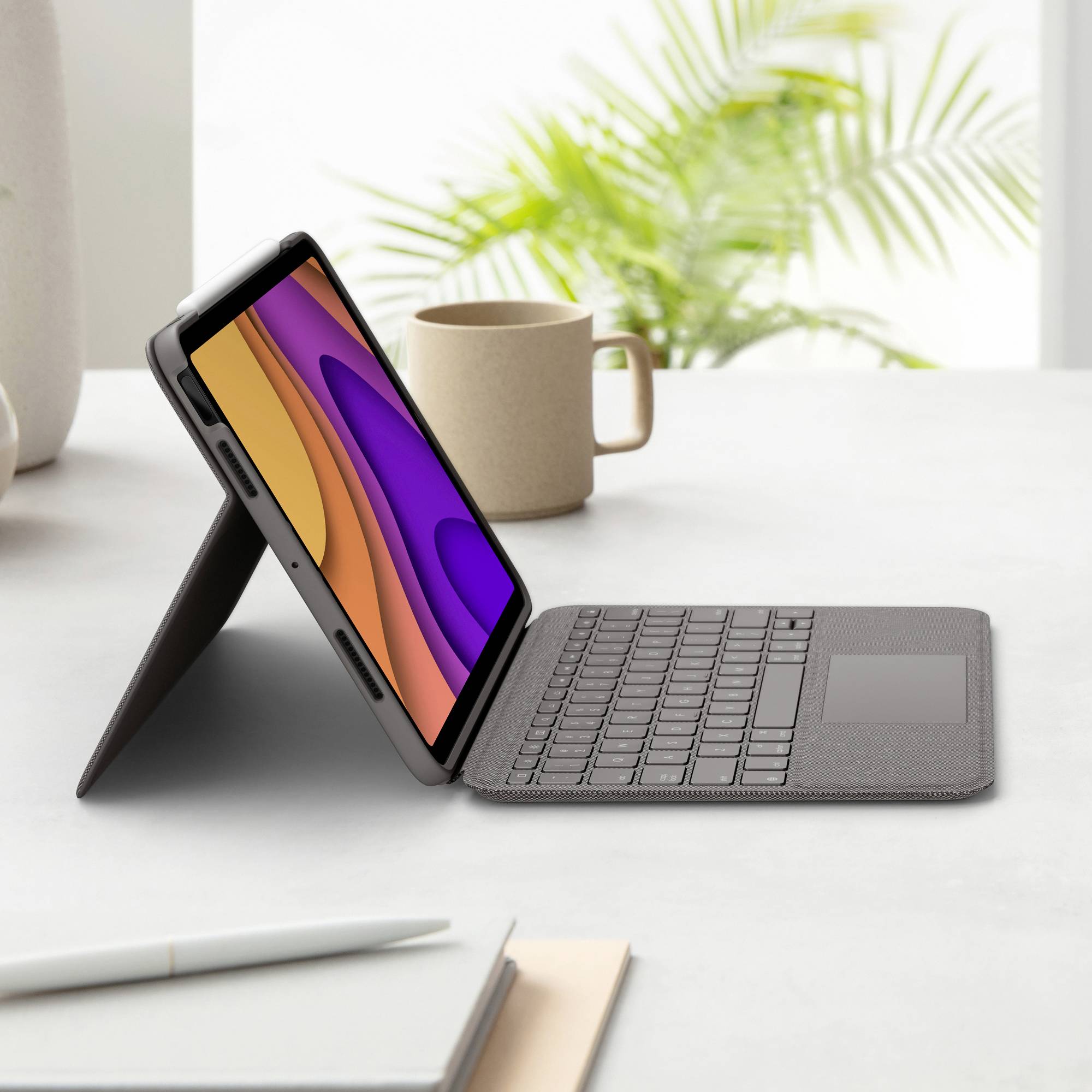 Tablet mit abnehmbarem Tastaturcover, im Profil auf Tisch stehend, Hintergrund mit verschwommener Pflanze und Tasse.