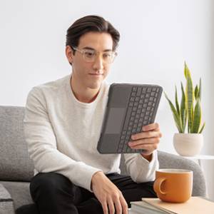 Eine Person in einem hellen Raum sitzt auf einem Sofa und hält ein Tablet mit Tastatur. Neben ihr steht eine orange Tasse und eine Pflanze.