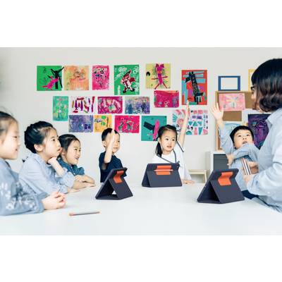 Eine Gruppe von Kindern sitzt an einem Tisch mit Tablets, einige heben die Hand. Eine Lehrerin sitzt rechts, bunte Bilder an der Wand.