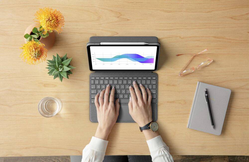 Eine Person tippt auf einer Tastatur, während ein Tablet einen farbigen Diagrammverlauf anzeigt, umgeben von Pflanzen und Arbeitszubehör.