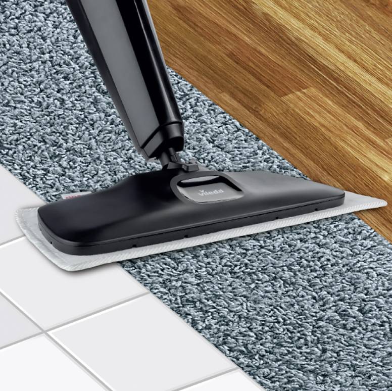 Ein Staubsauger reinigt einen Teppich am Rand eines Holzfußbodens neben weißen Fliesen, zeigt Bodenübergänge und Reinigungsfunktion.