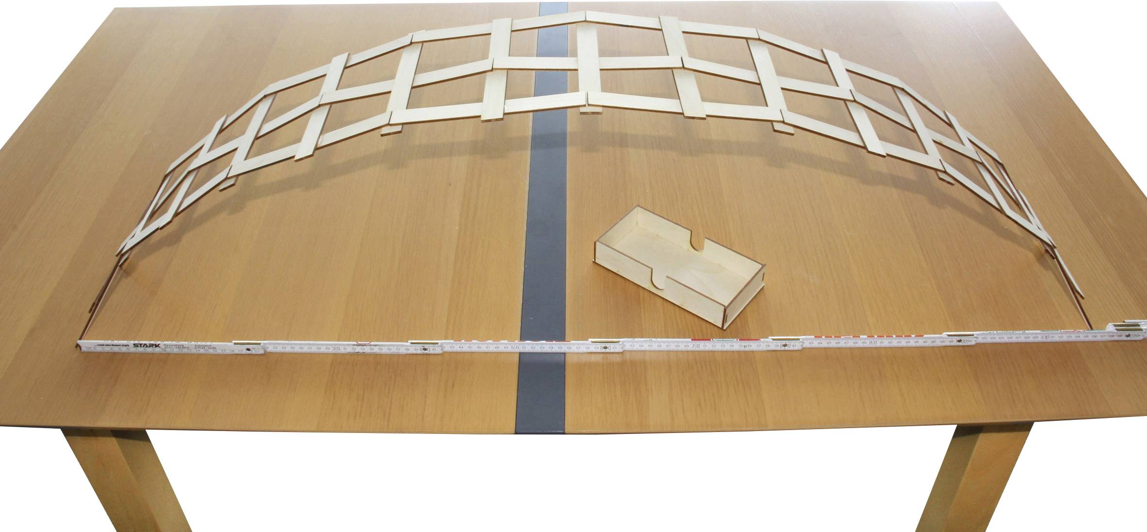 Ein Holzbogenmodell aus flachen Stäben liegt auf einem Tisch. Daneben ein kleines Holzkästchen. Ein Maßband erstreckt sich entlang des Bogens.
