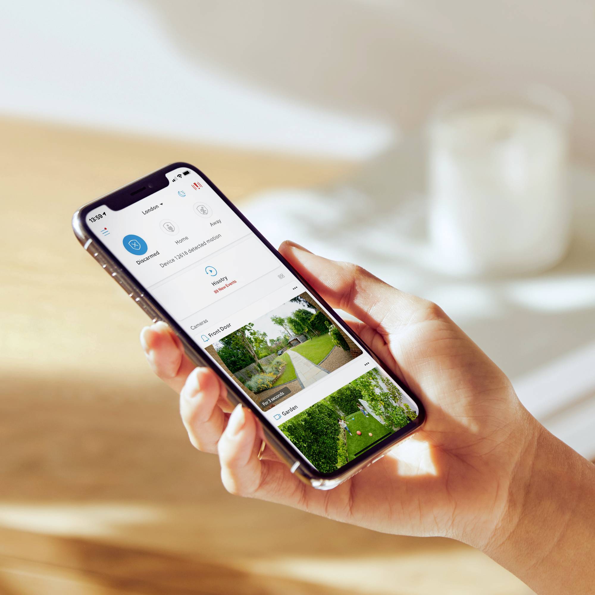 Eine Hand hält ein Smartphone, auf dem eine App geöffnet ist. Die App zeigt Kamerabilder und Optionen für Smart-Home-Geräte an.