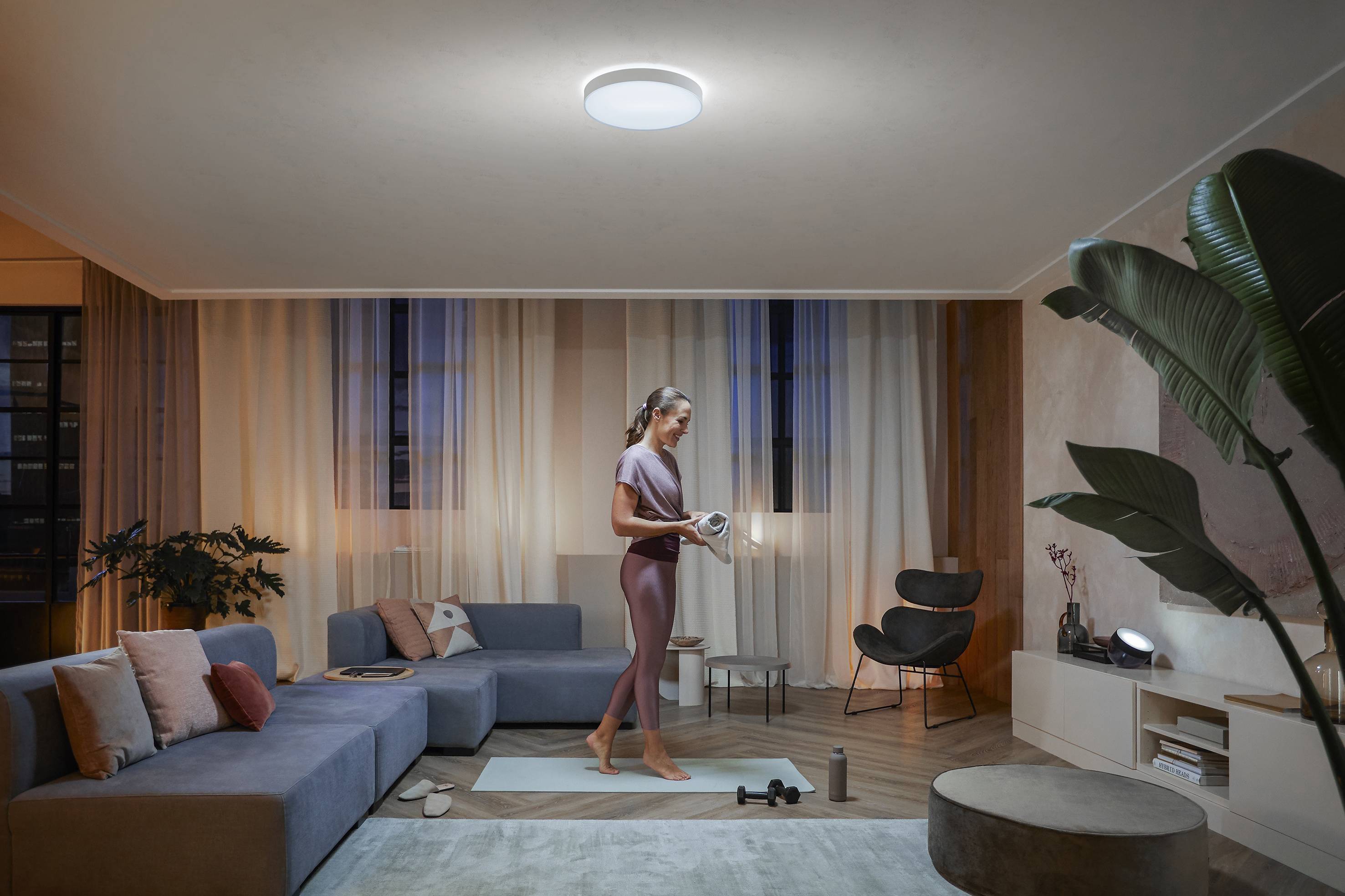 Eine Person macht Yoga in einem modernen, stilvoll eingerichteten Wohnzimmer mit einem großen Sofa und Pflanzen, Licht von der Deckenlampe.